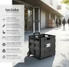 A black foldable shopping trolley with a telescopic handle is shown on a sidewalk in front of a building with people walking in the background. Náhled obrázku produktu