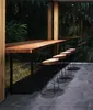 A wooden bar table with stools in an outdoor setting with lush greenery visible through a large window. Produktbild-Vorschau