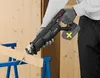 A person wearing gloves is using a cordless reciprocating saw to cut a piece of wood. The wood is supported by blue sawhorses. The background is a wooden wall. Produktbild-Vorschau