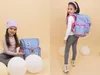 A split image showing two scenes of a young girl with a blue and pink backpack. The left side shows her sitting with the backpack next to her, and the right side shows her wearing the backpack. Produktbild-Vorschau