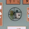 A round metal shelf mounted on a light green wall, decorated with a potted plant, a green glass, and a framed picture. Náhled obrázku produktu
