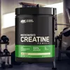 A container of creatine powder is shown in a gym setting. Produktbild-Vorschau