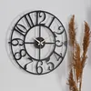 A black metal wall clock with Roman numerals is mounted on a light gray wall, with dried grass stalks to the right. Podgląd zdjęcia produktu
