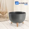 A dark gray ribbed planter pot with three wooden legs is displayed on a light wooden floor, with a decorative vase and a woven mat in the background. Náhled obrázku produktu
