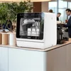 A compact dishwasher is shown on a countertop in a modern kitchen or office breakroom setting. There are plants and coffee-making equipment in the background. Produktbild-Vorschau