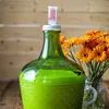 A green glass carboy with a fermentation airlock and orange gerbera daisies in a vase. Produktbild-Vorschau