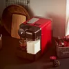 A red Tchibo Cafissimo coffee machine is shown in a kitchen setting, with coffee capsules in a wooden crate nearby. Produktbild-Vorschau