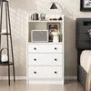 A white dresser with three drawers and an open shelf above, decorated with a speaker, a framed picture, and a vase of flowers. A bedside lamp and a floor lamp are also visible. Podgląd zdjęcia produktu