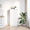 A white cabinet with two doors and a drawer is shown in a room next to a large green plant in a gray pot. To the left, a desk with drawers and a lamp is visible. There are also a few decorative items on top of the cabinet. Náhled obrázku produktu