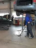 A mechanic is vacuuming up a spill in a garage while a car is lifted on a hydraulic lift. Podgląd zdjęcia produktu