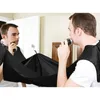 A man is shaving his beard in front of a mirror, using a black cape to catch the hair. Náhľad obrázku produktu