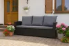A dark rattan sofa with grey cushions sits on a paved patio in front of a white house with wooden doors and shutters. Potted flowers in various colors surround the sofa. Anteprima dell’immagine prodotto