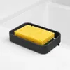 A black sponge holder with a yellow sponge inside, placed on the edge of a white sink. Náhled obrázku produktu