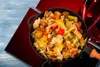 A close-up overhead view of a pan filled with stir-fried vegetables and chili peppers, placed on a blue wooden surface with red mats. Anteprima dell’immagine prodotto