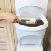 A hand is shown placing a glass bottle into a white trash can with a lid. Voir les images du produit