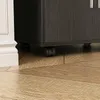 A close-up view of the bottom of a dark gray cabinet with black wheels, placed on a wooden floor next to a white baseboard. Náhled obrázku produktu
