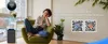 A woman is sitting on a chair and using her phone, with a device and a plant in the background. Produktbild-Vorschau