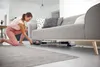 A woman is vacuuming under a sofa with a cordless vacuum cleaner. Produktbild-Vorschau