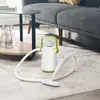 A portable steam cleaner is shown on a carpet in a living room setting, near a coffee table and a sofa. Náhľad obrázku produktu
