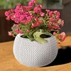 A white knitted-look planter with pink flowers sits on a wooden surface. Anteprima dell’immagine prodotto