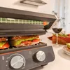 A close-up shot of a sandwich maker with sandwiches inside, set in a kitchen environment with a glass of wine and asparagus in the background. Voir les images du produit