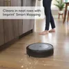 A robot vacuum cleaner is cleaning a wooden floor in a kitchen or living area, with a person's legs visible in the background. Voir les images du produit