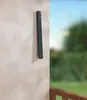 A dark gray metal bar is mounted vertically on a light beige wall, with a blurred background of green foliage and a blue sky visible to the right. A wooden railing is also visible in the lower right corner. Produktbild-Vorschau