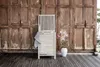 A wooden laundry basket with a striped towel draped over it, placed on a wooden deck in front of a rustic wooden door. A vase with pink flowers is to the right of the basket. Produktbild-Vorschau