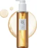 A bottle of cleansing oil with two droplets of oil next to it, set against a soft, blurred background. Anteprima dell’immagine prodotto