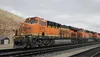A BNSF train with multiple locomotives on railroad tracks, with a building and hills in the background. Náhled obrázku produktu