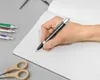 A hand holding a black pen and writing in a notebook, with other pens and scissors nearby. Produktbild-Vorschau