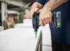 A person is using a cordless drill to screw into a white surface, likely in a workshop or construction setting. Produktbild-Vorschau