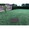 A circular black manhole cover is placed on a green lawn in a backyard. A wooden fence and trees are visible in the background. Podgląd zdjęcia produktu