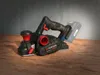A cordless planer sits on a wooden surface with a concrete wall in the background. Produktbild-Vorschau