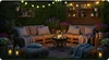 A cozy outdoor seating area at night, illuminated by string lights and a fire pit, with lush greenery in the background. Anteprima dell’immagine prodotto