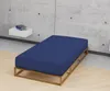 A dark blue fitted sheet on a wooden bed frame against a concrete wall. Produktbild-Vorschau