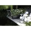 A dark planter filled with white flowers sits on a wooden deck next to two white chairs by a body of water. Podgląd zdjęcia produktu