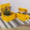 Two yellow wooden storage boxes are placed on a rug with a black and white geometric pattern. One box contains a potted lavender plant, and the other has books inside. The background is a brick wall. Produktbild-Vorschau