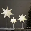 Three illuminated star lanterns of varying sizes are displayed in front of a dark wooden wall, with a Christmas tree visible on the right. Podgląd zdjęcia produktu