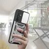 A hand holding a silver and black phone case with a ring holder, with a blurred background of a room. Produktbild-Vorschau