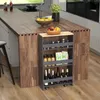 A narrow kitchen cart with four shelves is placed next to a wooden kitchen island. The cart is filled with various kitchen items like bottles, jars, and packets. The kitchen island has a sink, a bowl of fruit, and other kitchen accessories. Produktbild-Vorschau
