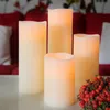 Four LED candles of varying heights are arranged on a white surface in front of a blurred red sofa with a floral patterned throw. Podgląd zdjęcia produktu