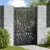 A black metal garden gate with a decorative cut-out pattern, set between two brick pillars and surrounded by greenery. Podgląd zdjęcia produktu