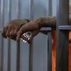 A close-up of a person's hands, with tattoos, holding a lock behind prison bars. Podgląd zdjęcia produktu