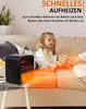 A child is sitting on a couch next to a small black heater that is emitting orange light. Produktbild-Vorschau