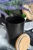 A black ceramic storage jar with a wooden lid is placed on a surface dusted with flour, with lavender and small potted plants in the background. Produktbild-Vorschau