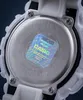 Close-up view of the back of a white Casio G-Shock watch, showing the metal case back with engraved text and a holographic authenticity sticker. Produktbild-Vorschau