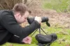 A man is lying on the grass, looking through a spotting scope on a tripod. Produktbild-Vorschau
