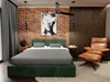 A bedroom scene featuring a green upholstered bed, a leather armchair with an ottoman, and brick and wood paneled walls. Produktbild-Vorschau