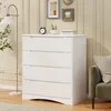 A white dresser with four drawers is shown in a room with a floor lamp and framed pictures on the wall. Náhled obrázku produktu